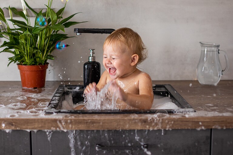 Cute happy baby girl with playing with water, soap bubbles and foam in a kitchen sink at home