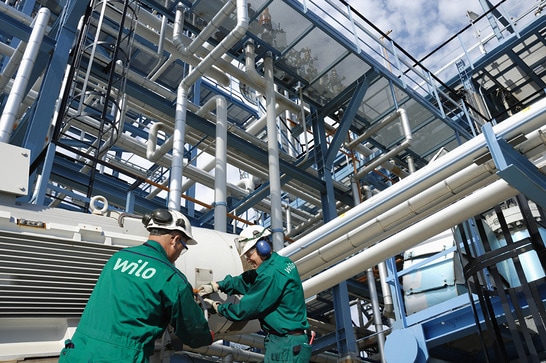 Industrial workers with green overalls and helmets are working in an industrial facility in front of pipes.