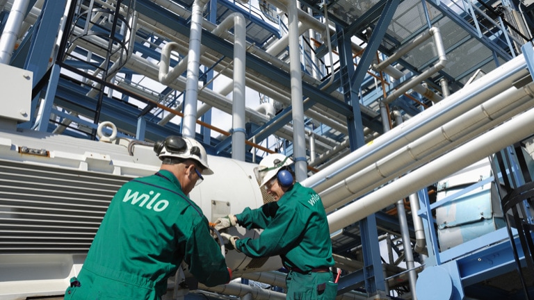 Industrial workers with green overalls and helmets are working in an industrial facility in front of pipes.