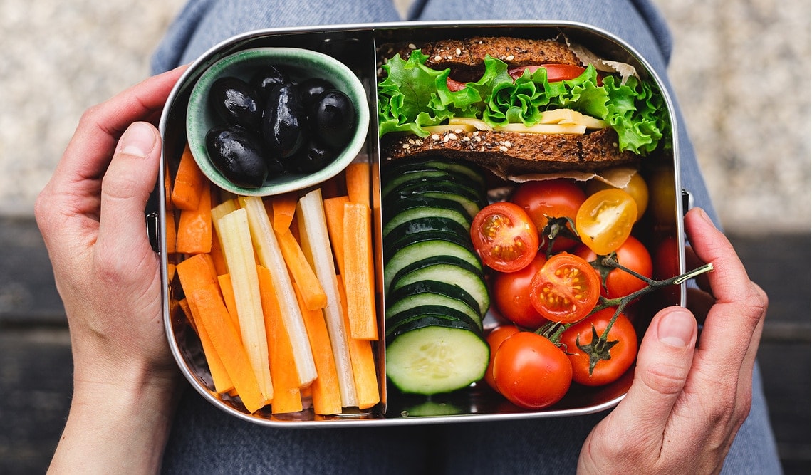 Point of view of a woman having a healthy lunch box. Black olives, carrots, cucumber, Cherry tomatoes and sandwich in the lunch box.