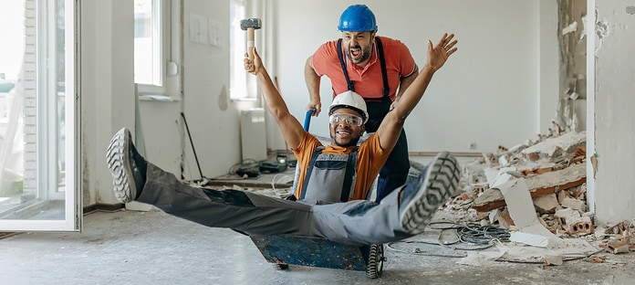 An African American male construction worker is smiling wide, while sitting on a construction trolley, pushed by his Caucasian coworker.