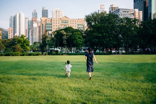 Rear view of happy young Asian mother and little daughter having fun outdoors, running around on lawn towards the city, enjoying Summer days in urban park on a lovely sunny day. Family lifestyle. Carefree and freedom concept. Connection to nature