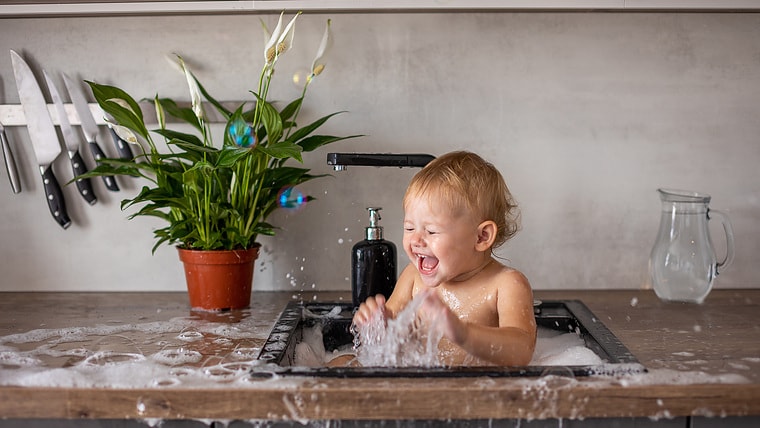 Cute happy baby girl with playing with water, soap bubbles and foam in a kitchen sink at home