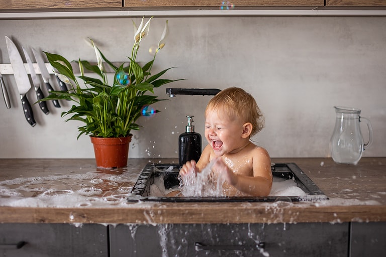 Cute happy baby girl with playing with water, soap bubbles and foam in a kitchen sink at home