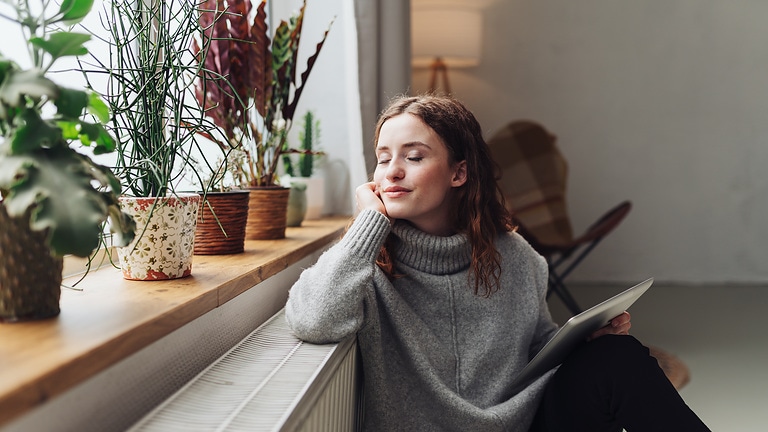 Eine entspannte Frau, die sehr zufrieden wirkt, sitzt mit einem Schreibblock und Wollpulli auf dem Boden vor der Heizung. Obwohl sie die Augen geschlossen hat, ist ihr Gesicht zum Fenster gerichtet. Auf der Fensterbank stehen viele Pflanzen.