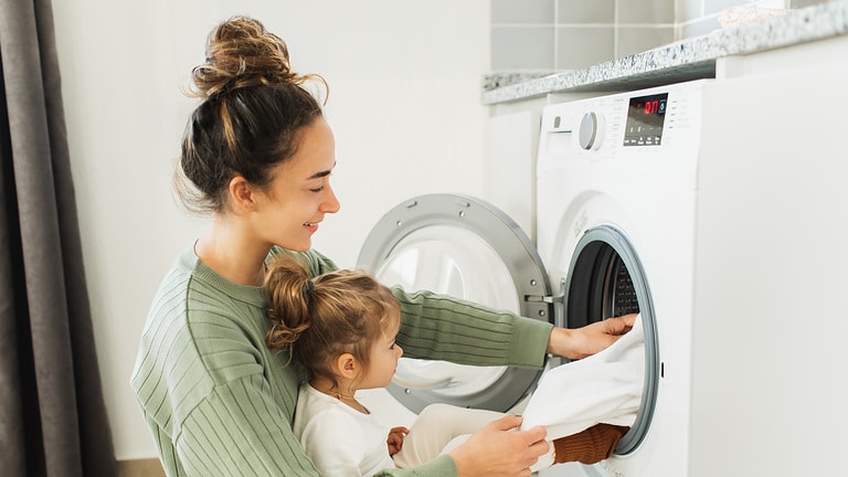 Eine Frau mit einem Kleinkind beschäftigt sich mit der Wäsche und faltet Kleidung in die Waschmaschine. Eine schöne Mutter und ihre Tochter haben Spaß und lachen, während sie die Wäsche in der Küche einer modernen Wohnung erledigen, in der eine Waschmaschine zu sehen ist.