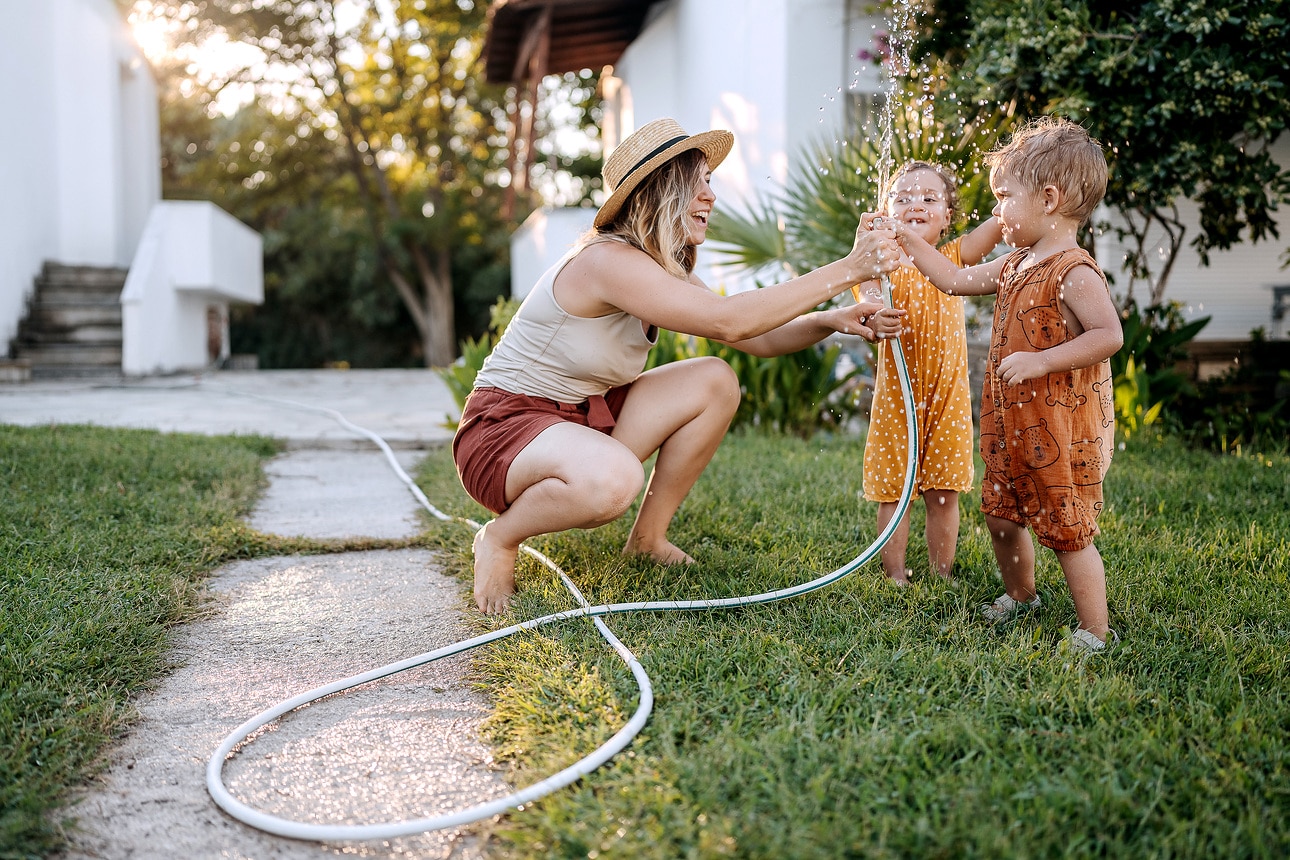 Mutter und ihre süßen kleinen Kinder spielen mit Wasser mit Gartenschlauch in einem Hinterhof