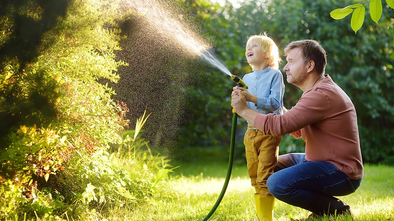 Ein Vater und sein Sohn stehen im sonnigen Garten, umgeben von blühenden Pflanzen und grünen Büschen. Gemeinsam halten sie den Gartenschlauch und bewässern die Pflanzen. Der Sohn steht zwischen den Beinen des Vaters, während beide den Wasserstrahl lenken. Beide wirken fröhlich und genießen die gemeinsame Zeit im Freien.