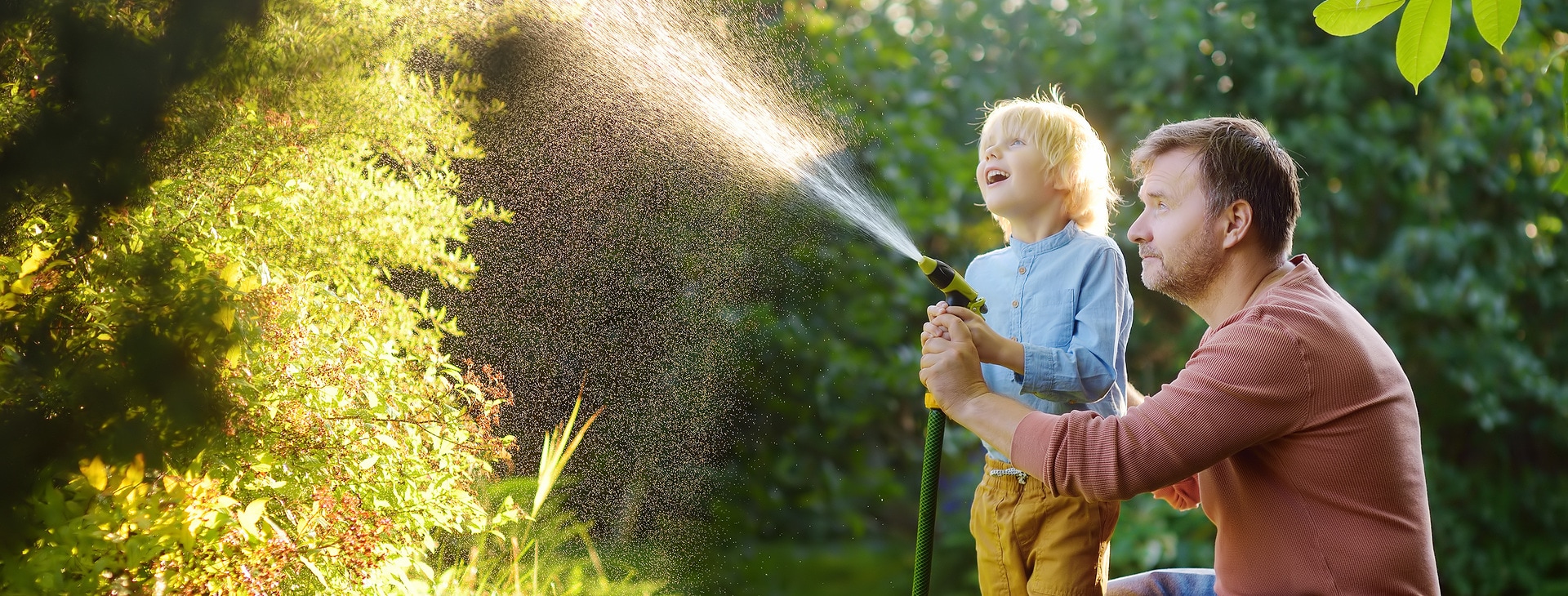 Ein Vater und sein Sohn stehen im sonnigen Garten, umgeben von blühenden Pflanzen und grünen Büschen. Gemeinsam halten sie den Gartenschlauch und bewässern die Pflanzen. Der Sohn steht zwischen den Beinen des Vaters, während beide den Wasserstrahl lenken. Beide wirken fröhlich und genießen die gemeinsame Zeit im Freien.
