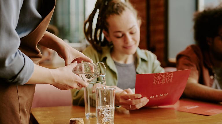 Klientka zamawiająca posiłek w punkcie gastronomicznym centrum handlowego oraz kelner nalewający świeżą, czystą wodę do picia.