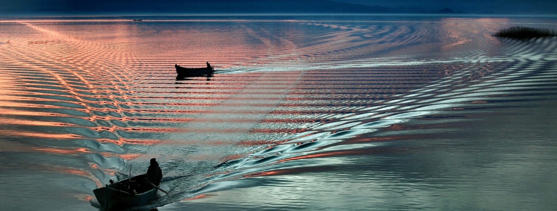 Two fishing boats on a lake in front of a mountain panorama at dusk.